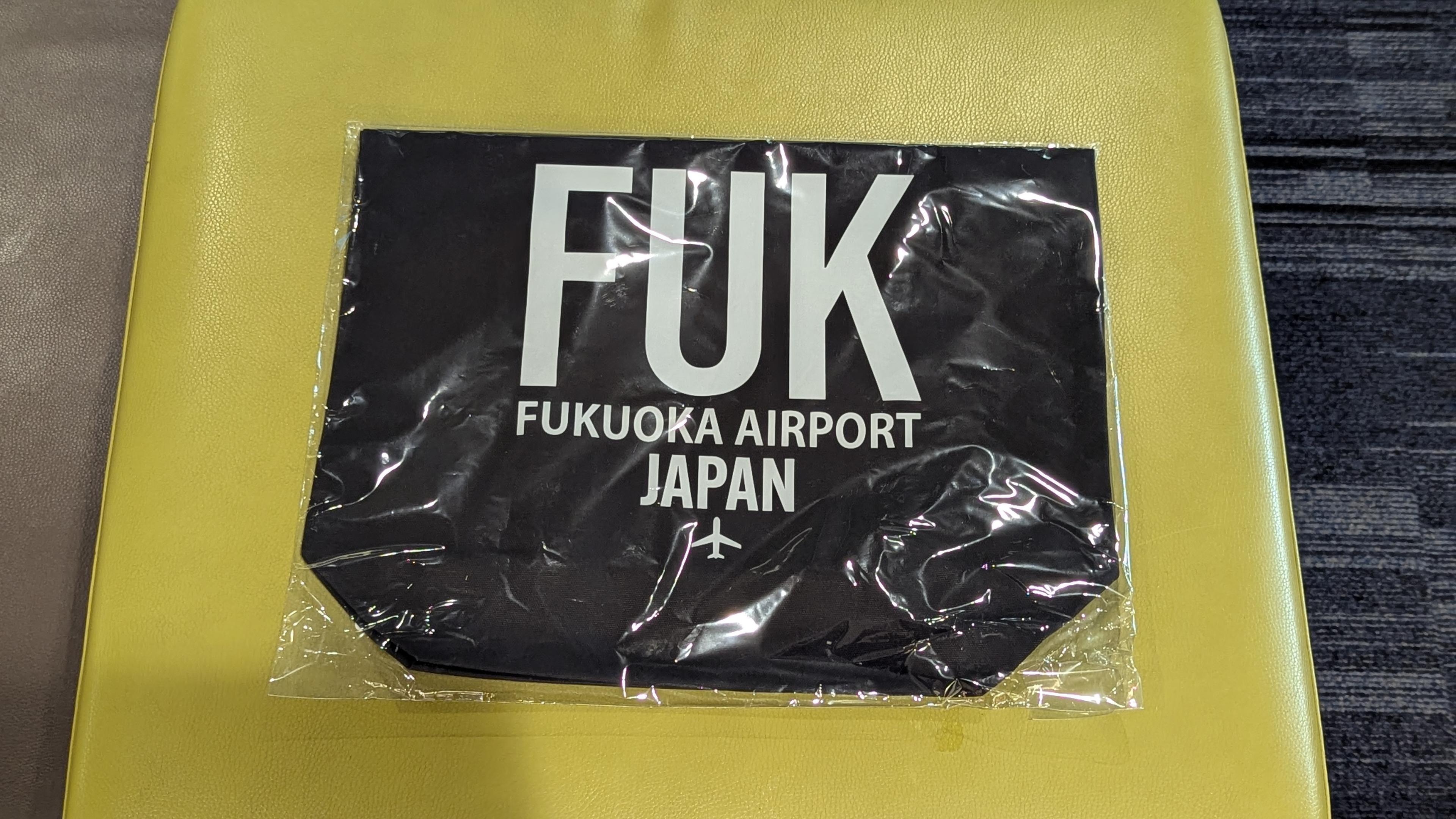 ビニール袋に入っている折りたたまれた福岡空港のスリーレタートートバッグ。全体は黒色で、文字は白になっている。FUKと大きく描かれていて下にはFUKUOKA AIRPORT JAPANと飛行機のマークが描かれている。